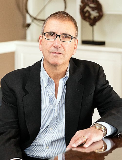 Professional headshot of Robert J. Gartland - middle-aged businessman wearing black business suit jacket over light blue and white striped dress shirt with open collar (no tie), short graying hair, black-rimmed glasses, positioned in confident pose with one hand resting on dark wooden surface in upscale office setting with white walls and built-in cabinetry in background