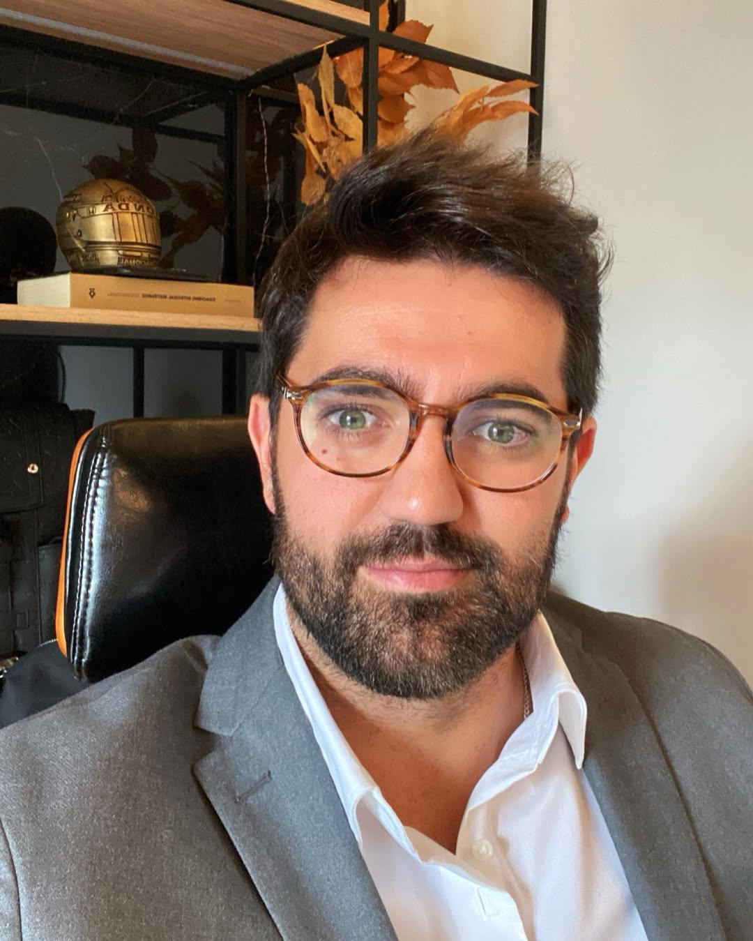 Professional headshot of Zandoriel Lubbert - man wearing glasses and a full beard, dressed in a gray suit jacket with white dress shirt, photographed in an office setting with shelves containing legal books and decorative items in the background