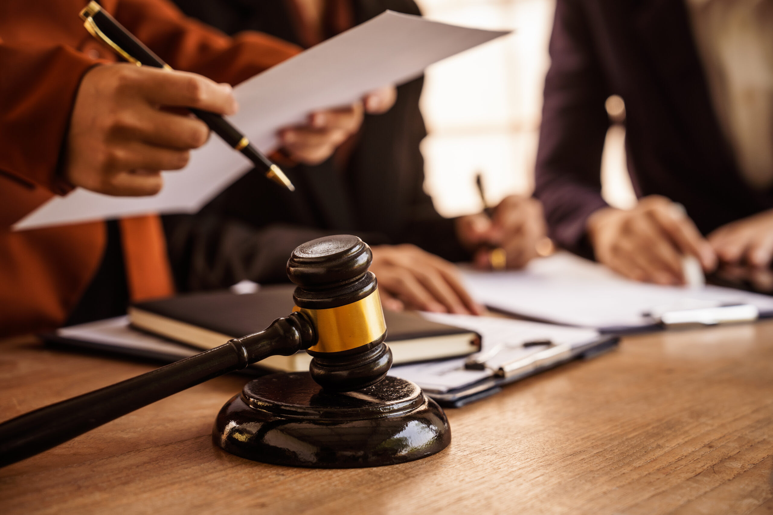 Professional legal office scene featuring a wooden judge's gavel with gold accent band prominently placed on a wooden desk in the foreground, with business professionals in suits reviewing legal documents and paperwork in the background